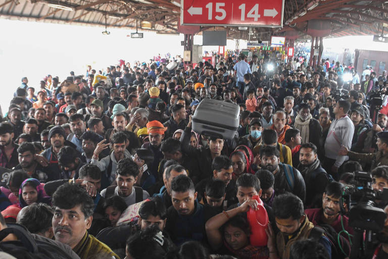 A imagem mostra uma grande multidão de pessoas em uma estação de trem， com muitos passageiros se aglomerando. Algumas pessoas estão segurando malas e outras estão usando máscaras. Ao fundo， há placas indicando os números 14 e 15. A atmosfera parece ser de movimento e expectativa.