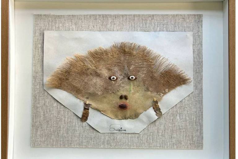 A imagem apresenta uma obra de arte emoldurada que utiliza elementos naturais para criar uma face. A face é composta por uma base de papel com texturas e cores neutras, e detalhes como pelos e olhos que se destacam. A obra é apresentada em uma moldura de madeira clara, com um fundo de tecido claro.