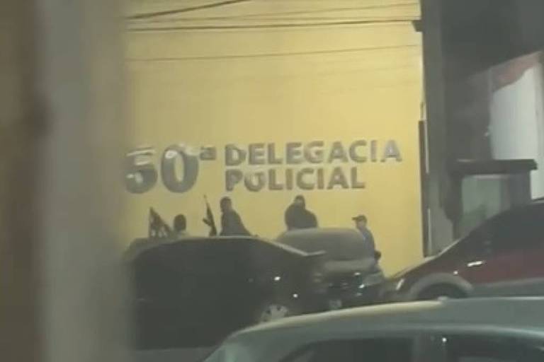 A imagem mostra uma parede amarela com a inscrição 039;50ª DELEGACIA POLICIAL039;. Algumas pessoas estão visíveis em frente à delegacia， e há veículos estacionados na parte inferior da imagem.