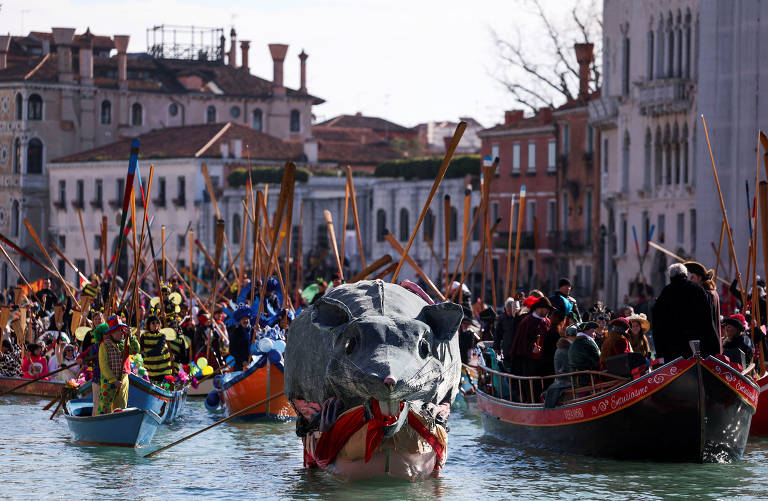 A imagem mostra uma cena do Carnaval de Veneza, com várias embarcações coloridas no canal. No centro, há uma grande figura de um rato, enquanto pessoas fantasiadas em trajes vibrantes e coloridos estão em diferentes barcos, cercadas por edifícios históricos ao fundo.