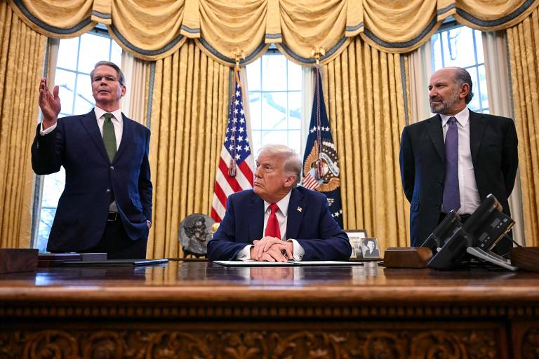 A imagem mostra uma reunião no Salão Oval da Casa Branca. Um homem de terno escuro e gravata verde está em pé à esquerda， gesticulando com a mão. No centro， um homem com cabelo claro e terno escuro está sentado à mesa， olhando para frente. À direita， um homem com cabelo grisalho e terno escuro está em pé， observando. Ao fundo， há cortinas amarelas e a bandeira dos Estados Unidos.