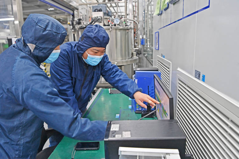 Dois trabalhadores usando roupas de proteção e máscaras estão em um ambiente industrial， observando e interagindo com um computador em uma mesa. O fundo mostra equipamentos industriais e tubulações.