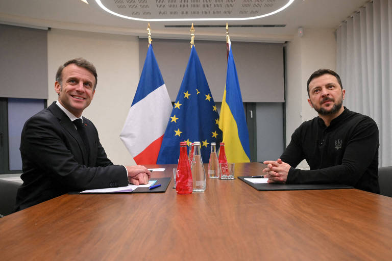 A imagem mostra dois homens sentados em uma mesa de madeira em uma sala de reuniões. À esquerda， um homem de terno escuro e gravata sorri， enquanto à direita， um homem com uma camisa preta e barba também sorri. Ao fundo， há três bandeiras: a da França， a da União Europeia e a da Ucrânia. Sobre a mesa， há garrafas de água e papéis.