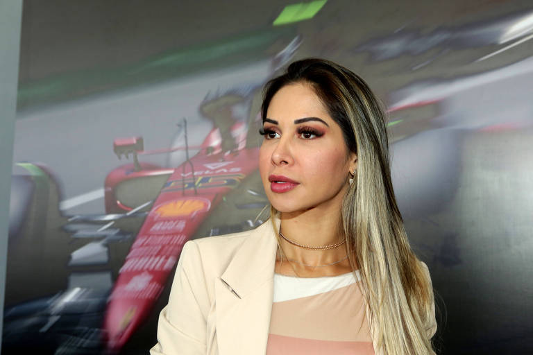 A imagem mostra uma mulher com cabelo longo e liso, usando um blazer claro sobre uma blusa de tom nude. Ela está em um evento relacionado à Fórmula 1, com um fundo desfocado que apresenta um carro de corrida vermelho. A mulher parece estar posando para a foto, com uma expressão séria.