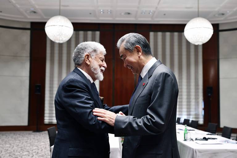 A imagem mostra dois homens em um ambiente de reunião. Eles estão se cumprimentando com sorrisos， um deles tem cabelo grisalho e barba， vestindo um terno escuro， enquanto o outro tem cabelo liso e escuro， usando um terno claro. Ao fundo， há uma mesa com cadeiras e uma decoração simples， com luzes pendentes no teto.