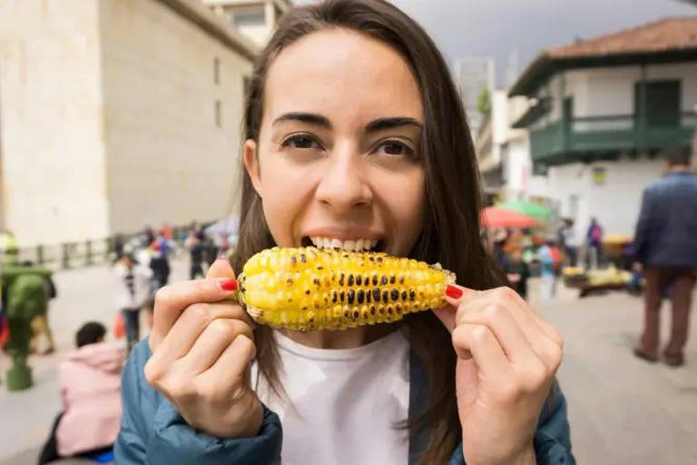 Uma mulher está segurando uma espiga de milho grelhado e sorrindo. Ela está em um ambiente ao ar livre， com várias pessoas ao fundo. A mulher tem cabelo longo e liso， e está vestindo uma jaqueta azul. O cenário parece ser uma praça ou mercado， com barracas e pessoas ao redor.