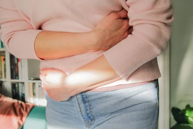 A imagem mostra uma pessoa de pé， usando um suéter rosa claro e jeans. A pessoa está segurando o abdômen com uma das mãos， enquanto a outra mão está apoiada no corpo. A luz do sol entra pela janela， iluminando parcialmente a cena. Ao fundo， há prateleiras com livros e uma planta.