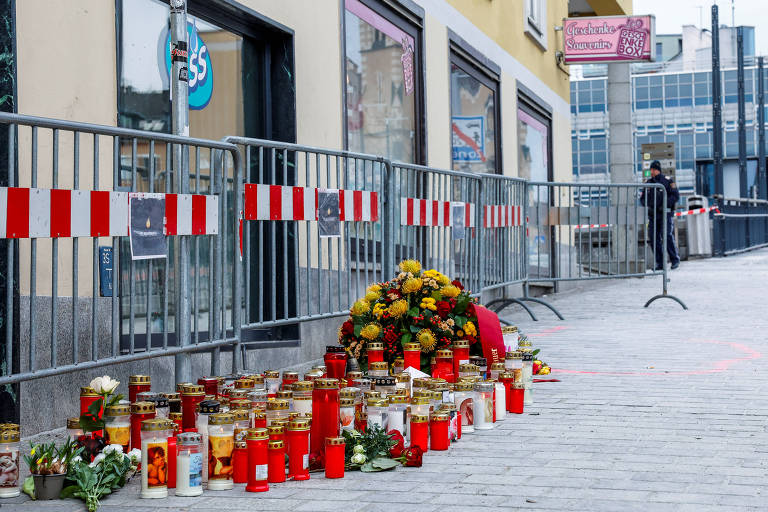 A imagem mostra uma área pública cercada por grades de segurança， onde há uma grande quantidade de velas acesas e flores dispostas em homenagem. As velas são de diferentes tamanhos e cores， com algumas sendo vermelhas e outras brancas. Ao fundo， há um comércio com janelas e uma placa visível， além de uma pessoa em pé， observando a cena. O ambiente parece ser urbano， com prédios e uma calçada pavimentada.