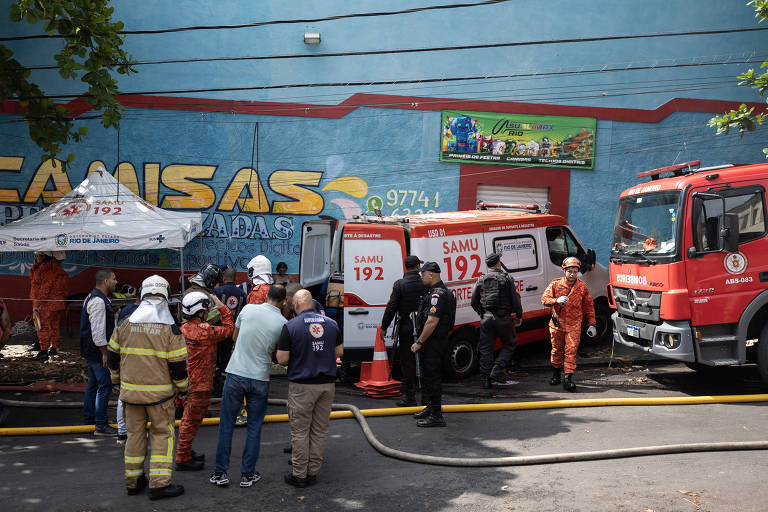 A imagem mostra uma cena de atendimento de emergência com várias pessoas e veículos de resgate. Há duas ambulâncias do SAMU 192 e um caminhão de bombeiros. Profissionais de resgate， incluindo bombeiros e policiais， estão reunidos em um local， enquanto algumas pessoas observam. Ao fundo， há uma parede azul com uma pintura e um banner visível.