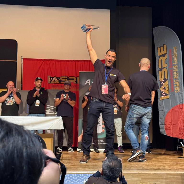 Um homem está em pé em um palco， levantando um troféu com a mão direita， enquanto sorri. Ele está vestido com uma camiseta preta e calças escuras. Ao fundo， há várias pessoas aplaudindo e um banner vermelho com a inscrição 039;WSRE039;. Outros participantes estão em pé， alguns de costas， e há uma mesa branca à frente.