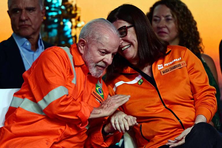 A imagem mostra duas pessoas em trajes de trabalho laranja， sorrindo e interagindo de forma amigável. Um homem com cabelo grisalho e uma mulher com cabelo escuro estão sentados próximos um do outro. Ao fundo， há outras pessoas assistindo ao evento， com um cenário que sugere um ambiente de celebração ou anúncio. O homem usa uma camisa com a bandeira do Brasil， enquanto a mulher tem um logotipo da Petrobras em sua roupa.