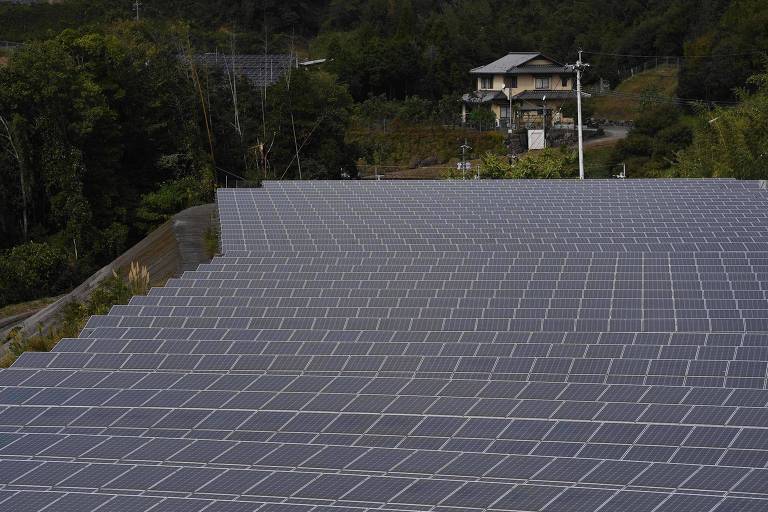 A imagem mostra uma grande instalação de painéis solares dispostos em fileiras em uma área rural. Ao fundo， há uma casa de dois andares e vegetação ao redor. A instalação solar ocupa a maior parte do primeiro plano， com os painéis refletindo a luz do sol.