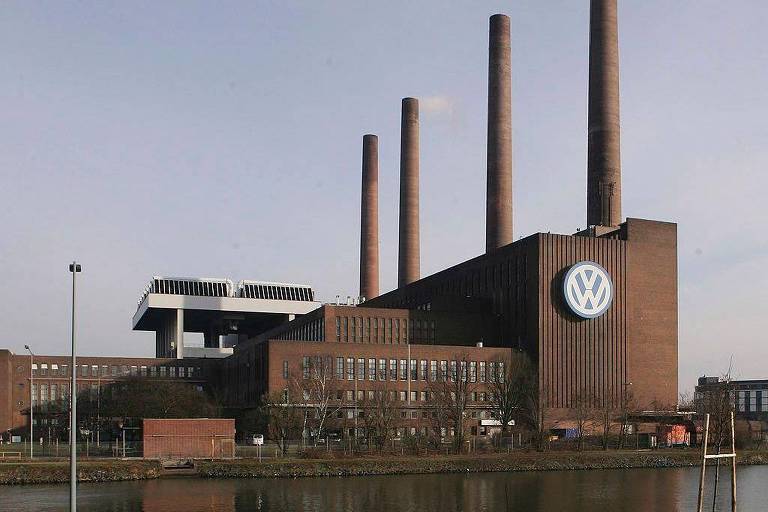 A imagem mostra a fábrica da Volkswagen， com uma estrutura de tijolos e quatro chaminés altas. O logotipo da Volkswagen é visível na parede do edifício. O céu está claro e a fábrica reflete na água em frente.