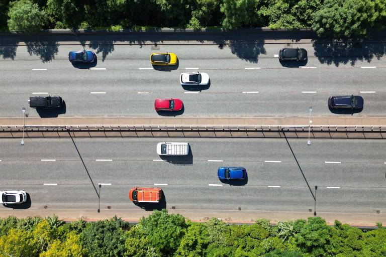 Imagem aérea de uma estrada com vários carros em movimento. Os veículos estão dispostos em duas faixas， com predominância de carros pretos， um carro vermelho， um amarelo e um branco. À direita da estrada， há uma faixa de vegetação densa.