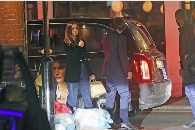 A imagem mostra uma cena noturna em uma rua, onde duas pessoas estão próximas a um carro preto. Uma mulher com cabelo solto e vestido escuro está de pé, parecendo estar em conversa ou esperando. Um homem, vestido com um terno escuro, está ao lado dela, enquanto uma mala e sacos de lixo estão no chão. Ao fundo, há luzes coloridas que iluminam a cena.