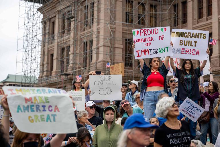 A imagem mostra um grupo de pessoas participando de um protesto em frente a um edifício. Algumas pessoas estão segurando cartazes com mensagens como 039;PROUD DAUGHTER OF AN IMMIGRANT039; e 039;IMMIGRANTS BUILT AMERICA039;. Outras mensagens visíveis incluem 039;IMMIGRANTS MAKE AMERICA GREAT039; e 039;STOP EION COUP039;. O ambiente é de mobilização， com bandeirinhas americanas e uma grande quantidade de pessoas reunidas.