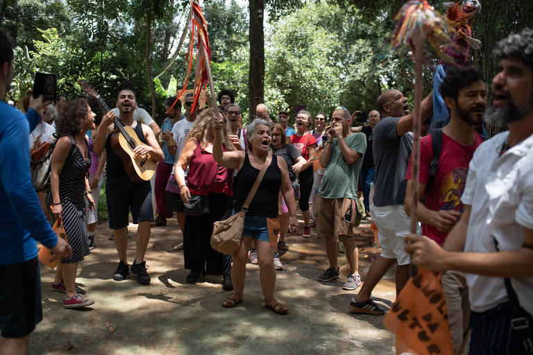 A imagem mostra um grupo de pessoas reunidas em um ambiente ao ar livre， cercado por árvores. Algumas pessoas estão dançando e cantando， enquanto outras seguram instrumentos musicais e faixas. A atmosfera é de celebração e união， com expressões de alegria e entusiasmo entre os participantes.