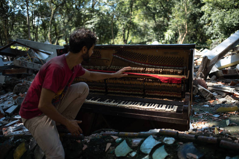 Um homem está sentado em um ambiente ao ar livre， cercado por destroços e vegetação. Ele está interagindo com um piano antigo， que está parcialmente exposto e danificado. O piano tem algumas cordas visíveis e uma parte interna exposta. O cenário é de um local abandonado， com árvores ao fundo e detritos ao redor.