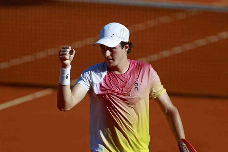 Um jogador de tênis está em uma quadra de saibro， levantando o punho em sinal de vitória. Ele usa uma camiseta com um gradiente de cores que vai do rosa ao amarelo e um boné branco. O fundo mostra a rede da quadra de tênis.