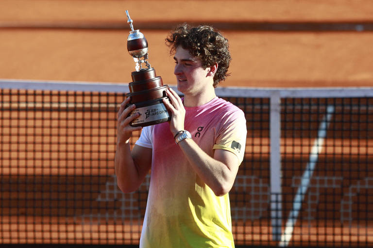 Um jovem jogador de tênis está segurando um troféu em uma quadra de saibro. Ele está vestindo uma camiseta com gradiente de cores que vai do rosa ao amarelo. O fundo mostra a rede da quadra e a superfície de terra batida.