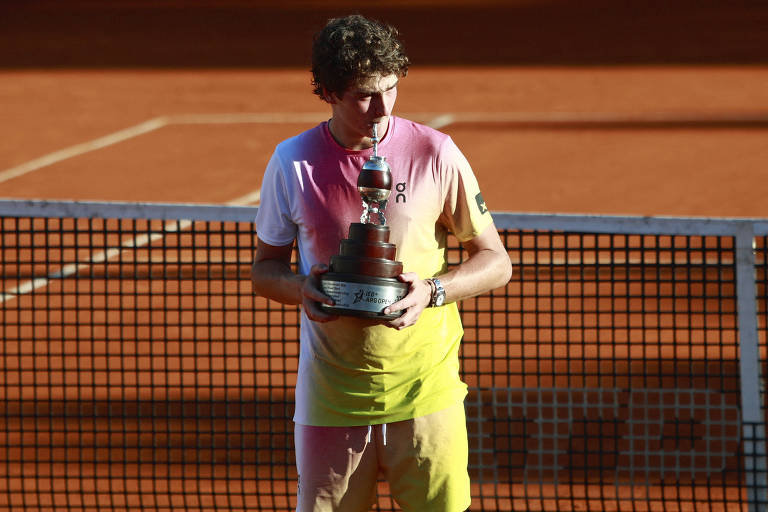 Um jogador de tênis está segurando um troféu em uma quadra de saibro. Ele usa uma camiseta com cores que vão do branco ao amarelo e shorts amarelos. O fundo mostra a rede da quadra e a superfície de saibro.