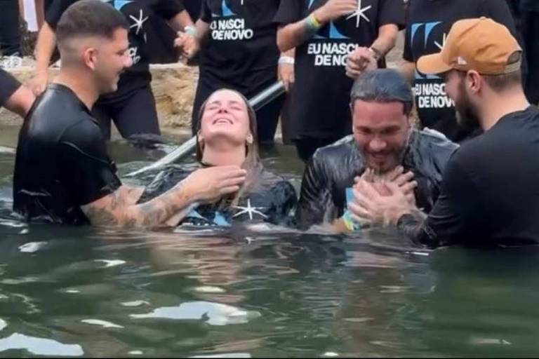 A imagem mostra um grupo de pessoas em um local aquático, onde algumas estão sendo batizadas. Duas pessoas estão parcialmente submersas na água, enquanto outras as cercam, demonstrando apoio e alegria. Todos estão vestindo camisetas pretas com a frase 'Eu Nasci de Novo' visível. As expressões faciais refletem emoção e felicidade.