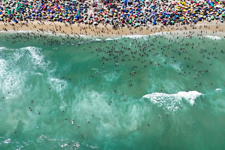 A imagem mostra uma vista aérea de uma praia lotada, com muitas pessoas na água e na areia. Há uma grande quantidade de guarda-sóis coloridos dispostos na areia, criando um padrão vibrante. As ondas do mar estão visíveis, com algumas pessoas nadando e outras caminhando na água.