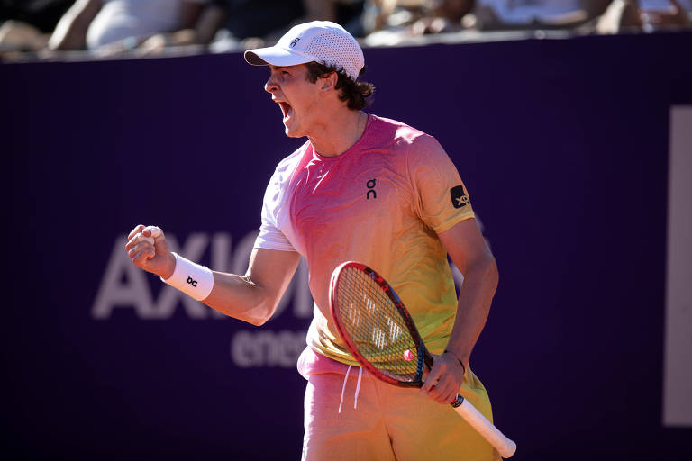 Um jogador de tênis está comemorando com um grito de vitória， segurando uma raquete. Ele usa uma camiseta de cores gradientes， que vão do rosa ao amarelo， e um boné branco. Ao fundo， é possível ver uma arquibancada com espectadores.