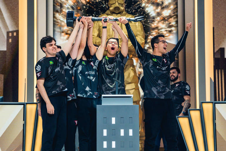 Quatro jogadores de eSports estão levantando um troféu em um palco， comemorando uma vitória. Eles estão vestidos com camisetas escuras e parecem animados. Ao fundo， há uma estátua dourada e efeitos de luz que criam um ambiente festivo.