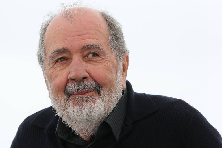 Um homem idoso com barba e cabelo grisalho， vestindo uma camisa escura e um suéter preto. Ele está sorrindo levemente e olhando para a câmera， com um fundo claro e desfocado.