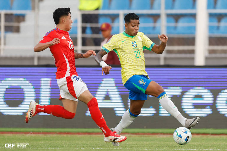 A imagem mostra um momento de uma partida de futebol entre duas equipes. Um jogador está vestindo uma camisa amarela com detalhes em verde e calças brancas， enquanto o outro jogador está usando uma camisa vermelha e calças brancas. O jogador da camisa amarela está em movimento， tentando chutar a bola， enquanto o jogador da camisa vermelha se aproxima para defender. O campo de futebol é visível ao fundo， com arquibancadas parcialmente ocupadas.