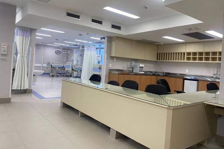 A imagem mostra o interior de um hospital， com uma área de recepção ou enfermagem à frente， onde há uma mesa longa com cadeiras pretas. Ao fundo， é possível ver várias camas hospitalares separadas por cortinas brancas. O ambiente é bem iluminado， com paredes em tons claros e armários na parte superior da mesa.