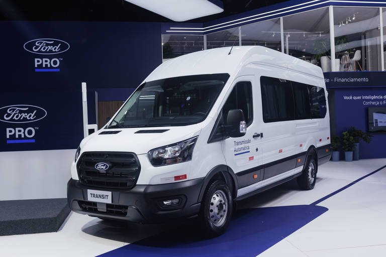 A imagem mostra um furgão Ford Transit， de cor branca， estacionado em um estande. O veículo possui janelas grandes e um design moderno， com o logotipo da Ford visível na frente. O fundo apresenta um ambiente de exposição com elementos de marketing da Ford.