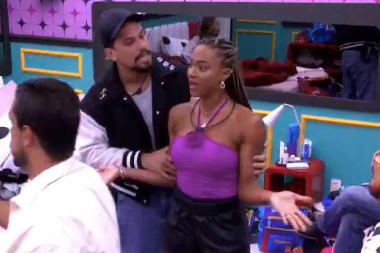 A imagem mostra um grupo de pessoas em um ambiente interno colorido. Um homem está segurando o braço de uma mulher, que parece estar expressando surpresa ou indignação. A mulher usa uma blusa roxa e calças escuras, enquanto o homem veste uma jaqueta preta. Ao fundo, outras pessoas estão sentadas e interagindo, com móveis e objetos visíveis na sala.