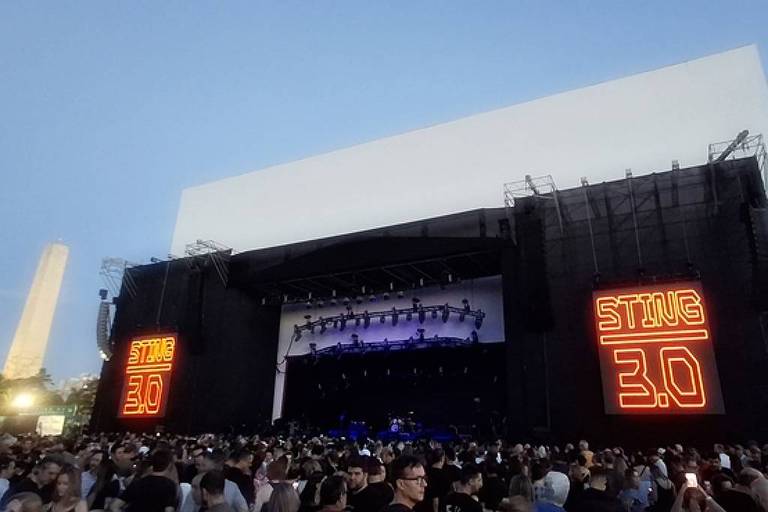 A imagem mostra um grande palco de show com uma multidão de pessoas na frente. À esquerda do palco， há um grande letreiro iluminado com a palavra 039;STING039; e o número 039;3.0039;. O céu está claro， indicando que o evento ocorre ao entardecer. Ao fundo， é possível ver um monumento ou obelisco.