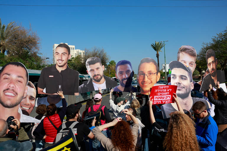 A imagem mostra um grupo de pessoas participando de um protesto. Elas seguram grandes cartazes com rostos de diferentes indivíduos. Algumas pessoas estão vestindo camisetas escuras e outras têm cabelos longos e cacheados. Ao fundo, há árvores e edifícios. Um dos cartazes contém texto em hebraico, que parece ser uma mensagem de protesto.