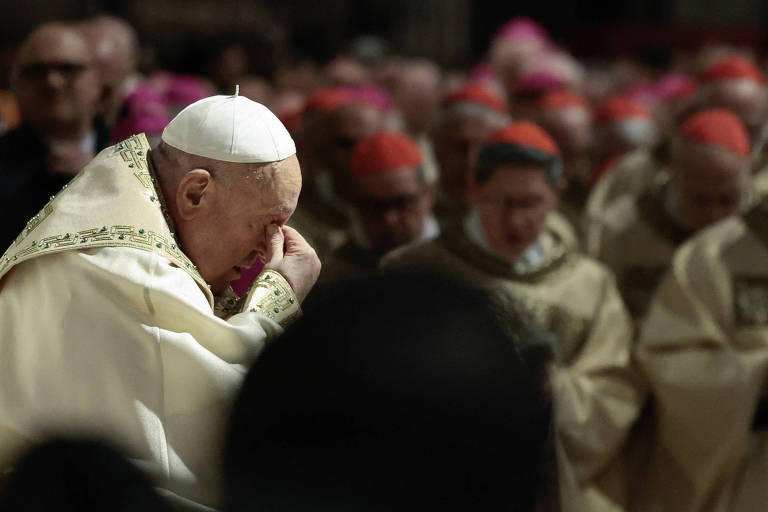 A imagem mostra o Papa Francisco， com vestes brancas e douradas， em um momento de reflexão ou oração. Ele está com a cabeça inclinada e uma mão no rosto， enquanto cardeais ao fundo estão em posição de oração， usando vestes semelhantes. O ambiente parece ser uma catedral， com uma iluminação suave.