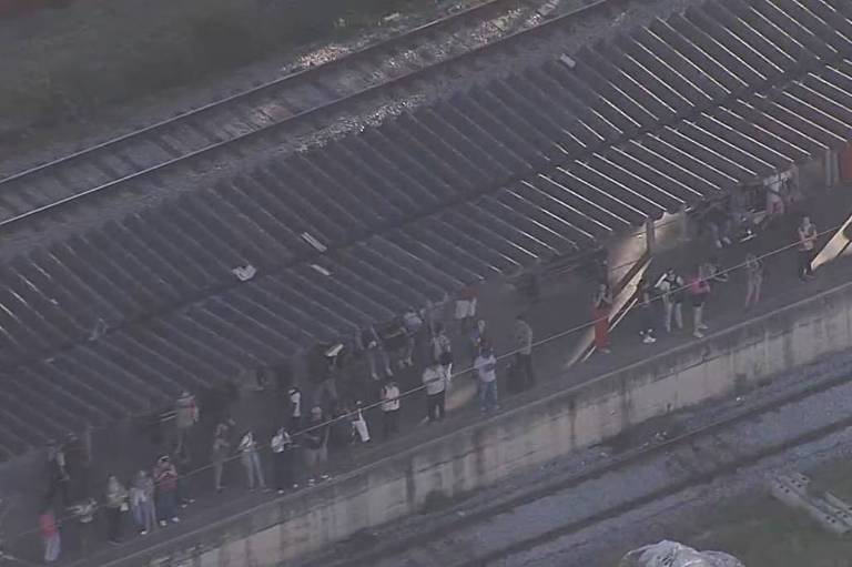 Imagem aérea de uma estação de trem onde várias pessoas estão esperando. A plataforma é coberta e há trilhos de trem visíveis ao lado. O ambiente parece ser ao ar livre， com vegetação ao fundo.