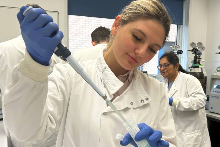 Uma mulher está usando uma micropipeta em um laboratório. Ela está vestindo um jaleco branco e luvas azuis， concentrada na tarefa. Ao fundo， outras pessoas também estão trabalhando em um ambiente de laboratório， com equipamentos visíveis como cilindros e uma máquina. A iluminação é clara e o ambiente parece organizado.