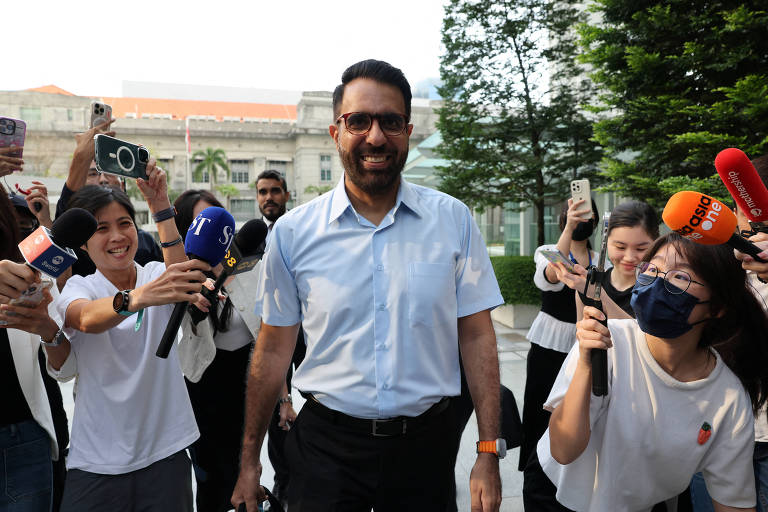 Um homem sorridente， usando uma camisa azul clara e óculos， caminha em direção à câmera enquanto é cercado por um grupo de pessoas. Algumas delas seguram microfones e celulares， tentando registrar o momento. O ambiente é ao ar livre， com árvores e um edifício ao fundo.