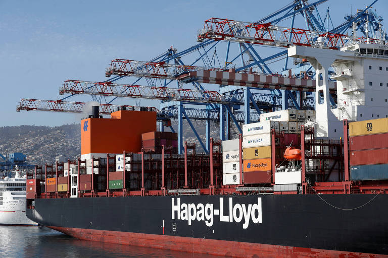 A imagem mostra um navio de carga da empresa Hapag-Lloyd atracado em um porto. O navio é carregado com contêineres de diferentes cores， incluindo laranja e verde. Ao fundo， há guindastes de carga azuis， utilizados para movimentar os contêineres. O céu está claro e o ambiente parece ser um porto movimentado.