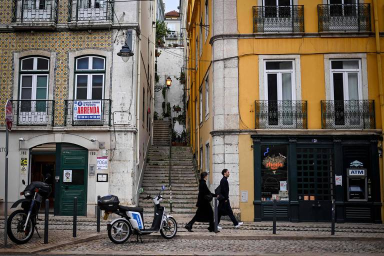 A imagem mostra uma rua em Lisboa com edifícios de cores diferentes. À esquerda， um prédio com azulejos e uma placa de 039;Arrenda039; visível. À direita， um prédio amarelo com janelas e varandas. No centro， uma escadaria que leva a um nível superior. Duas pessoas caminham pela calçada， uma delas vestindo roupas escuras. Há uma motocicleta estacionada na calçada e um caixa eletrônico visível na fachada do prédio à direita.