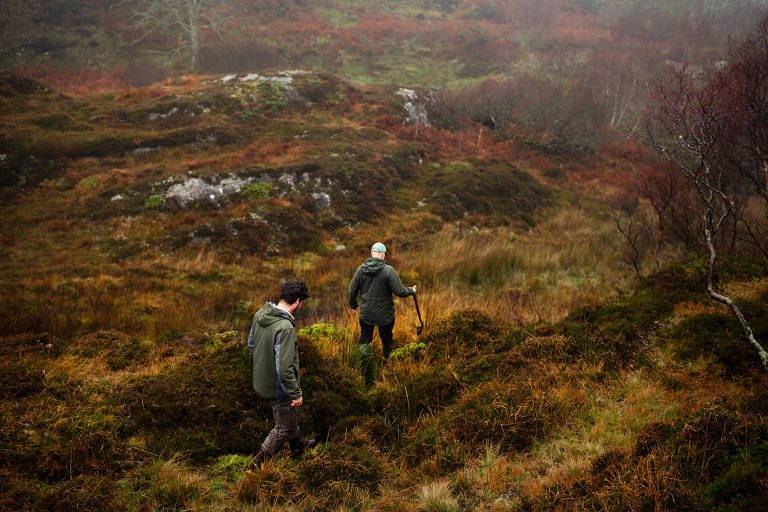 A imagem mostra duas pessoas caminhando em um terreno acidentado e coberto de vegetação. O ambiente é nebuloso， com árvores esparsas e rochas visíveis ao fundo. As pessoas estão vestidas com roupas de inverno e parecem estar explorando a área.