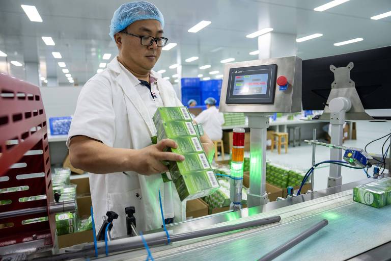 Um trabalhador em uma fábrica está segurando várias caixas verdes enquanto se prepara para colocá-las em uma esteira. Ele usa um jaleco branco e uma touca azul. Ao fundo， há outros trabalhadores e uma máquina com um painel digital que exibe luzes e botões.