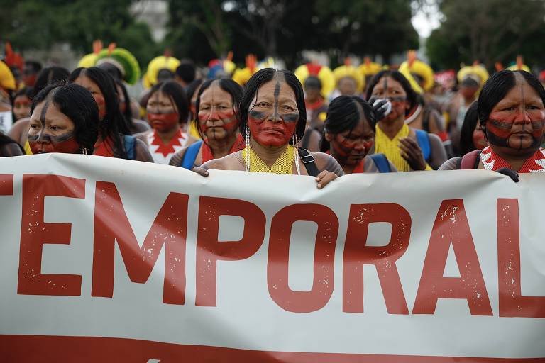 A imagem mostra um grupo de pessoas indígenas marchando em uma manifestação. Elas têm o rosto pintado com tinta vermelha e usam roupas tradicionais. Algumas pessoas estão segurando uma faixa grande que contém a palavra 039;TEMPORAL039; em letras grandes e vermelhas. Ao fundo， há outras pessoas e elementos da natureza.