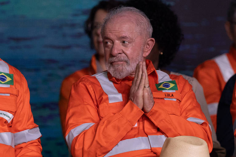 Um homem com cabelo grisalho e barba， vestindo uma camisa de trabalho laranja com faixas refletivas brancas. Ele está sentado， com as mãos unidas em um gesto de oração ou agradecimento， e parece estar em um evento. Ao fundo， há outras pessoas e um cenário que sugere um ambiente de trabalho ou uma cerimônia.