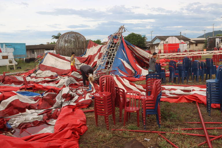 A imagem mostra um espaço ao ar livre onde um circo foi desmontado. Há uma grande lona de circo vermelha e branca caída no chão， com algumas partes amontoadas. Várias cadeiras plásticas vermelhas estão espalhadas pelo local. Duas pessoas estão trabalhando na área， uma delas está em pé e a outra está agachada， aparentemente organizando a lona. Ao fundo， há uma estrutura circular que parece ser uma parte do circo， além de algumas construções e um céu nublado.