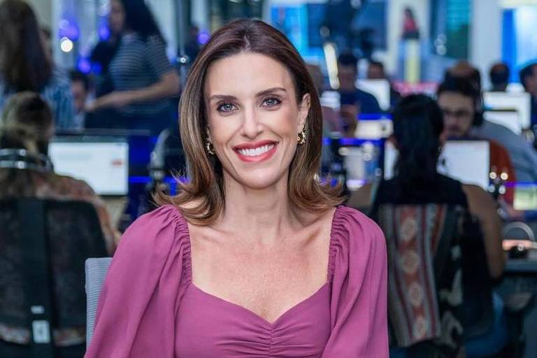 Uma mulher sorridente com cabelo castanho claro e liso, vestindo uma blusa roxa com mangas bufantes, está sentada em um estúdio de televisão. Ao fundo, várias pessoas estão trabalhando em computadores, com telas visíveis. A iluminação é suave e o ambiente parece moderno.