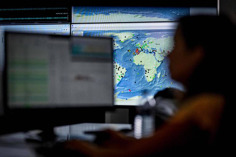 A imagem mostra uma pessoa em foco desfocado， sentada em frente a um computador. Ao fundo， há três monitores exibindo um mapa-múndi com várias marcações coloridas， possivelmente indicando locais de interesse geográfico ou eventos. O ambiente parece ser um escritório ou sala de controle.