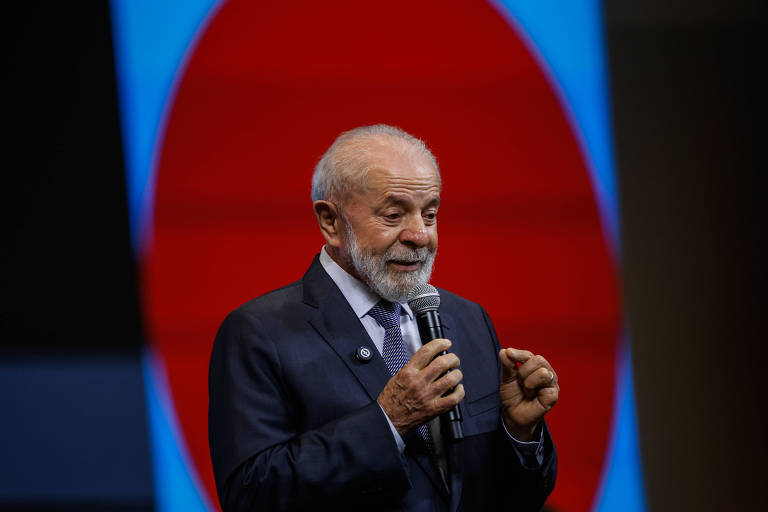 Um homem de cabelo grisalho e barba, vestido com um terno escuro e gravata, está falando ao microfone em um palco. Ao fundo, há um grande painel com um círculo vermelho e um fundo azul. O homem parece estar gesticulando enquanto se dirige ao público.